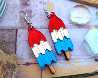May include: A pair of dangle earrings shaped like rocket popsicles. Each earring features three stacked layers of red, white, and blue acrylic with a small brown wooden stick at the bottom. The earrings hang from silver-toned metal fishhook wires.