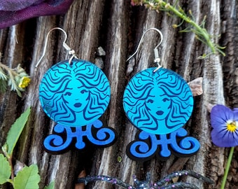 May include: A pair of dangle earrings featuring a circular bright blue acrylic charm with a black line art illustration of a face with flowing hair, attached to a black scrollwork base with silver-toned metal earring hooks for pierced ears.