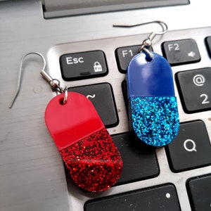 May include: Two novelty earrings shaped like capsules. One is red with red glitter, the other is blue with blue glitter. Both have silver-colored ear wires and are displayed on a laptop keyboard.