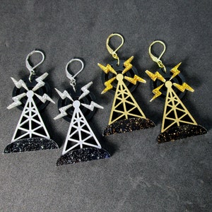 Pode incluir: Três pares de brincos com um design de torre de rádio preta e prateada com raios. Os brincos são feitos de metal e têm um acabamento brilhante.