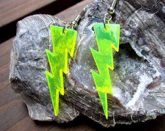 May include: A pair of neon green lightning bolt earrings. The earrings are made of a translucent material and have a jagged, zig-zag design. They hang from silver-colored fish hook ear wires. The earrings are displayed on a gray rock surface.