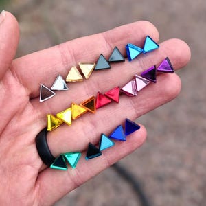Könnte beinhalten: Eine Sammlung kleiner, dreieckiger Ohrstecker in einer Vielzahl von reflektierenden, metallischen und leuchtenden Farben, darunter Gold, Silber, Schwarz, Blau, Rot, Violett und Türkis, die in Reihen auf der Handfläche angeordnet sind.