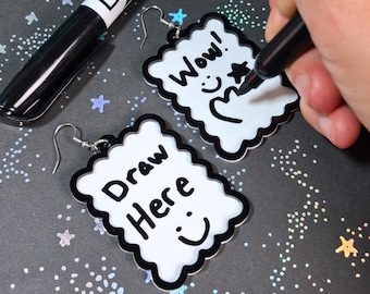 May include: Two rectangular white earrings with scalloped black edges and silver-toned hooks. One earring features the text "Draw Here" with a smiley face, while the other displays "Wow!" with a star and a hand-drawn heart shape in black ink.