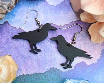 May include: A pair of black raven-shaped earrings with silver-colored hooks. The earrings are displayed on a colorful background with flowers and crystals. The raven earrings are a unique accessory.