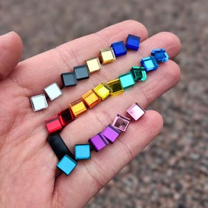 May include: A collection of small, square, reflective mirror tiles in a variety of colors including red, orange, yellow, green, blue, purple, pink, silver, gold, and black. These tiny, flat, geometric pieces have a smooth, glossy finish.