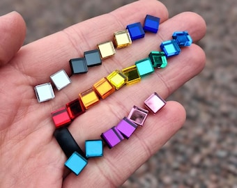 May include: A collection of small, square, reflective mirror tiles in a variety of colors including red, orange, yellow, green, blue, purple, pink, silver, gold, and black. These tiny, flat, geometric pieces have a smooth, glossy finish.