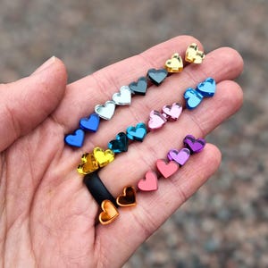 May include: A collection of small, heart-shaped studs in a variety of vibrant colors including gold, silver, blue, pink, purple, orange, and red. Each heart features a smooth, reflective surface and is arranged in rows across the palm of a hand.