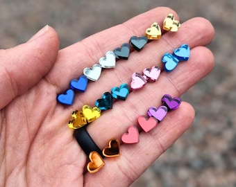May include: A collection of small, heart-shaped studs in a variety of vibrant colors including gold, silver, blue, pink, purple, orange, and red. Each heart features a smooth, reflective surface and is arranged in rows across the palm of a hand.