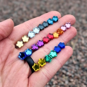 May include: A collection of small, flower-shaped acrylic beads in a variety of vibrant colors including blue, green, yellow, red, purple, pink, orange, silver, and gold, arranged in rows on a flat surface with a black ring base.