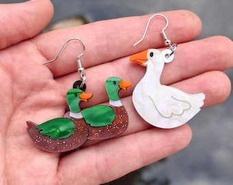 May include: A pair of dangle earrings featuring acrylic charms. One earring depicts a mallard duck with a green head, brown body, and orange beak. The other earring shows a white goose with an orange beak. Both earrings have silver-toned hooks.