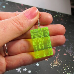 May include: A neon green acrylic earring with a silver hook. The earring features a rectangular shape with a grid of letters and numbers printed on it. The text on the earring reads: "OLE20989 Riovoip uk ?26 x7 W5944? 15 QU 136K ?1 87 105 31 VOR 191 SD 4 Jer 41 G"