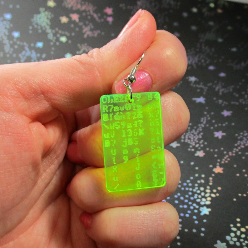 May include: A neon green acrylic earring with a silver hook. The earring is rectangular and has a grid of letters and numbers printed on it. The text on the earring reads: "0120984 @726? W594? 5 QU 136K 71 87 385 51 Uon SD 91 X JC U 11"