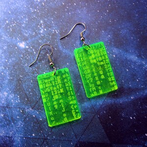 May include: A pair of green acrylic earrings with silver hooks. Each earring features a rectangular shape with a random assortment of letters and numbers printed on it. The earrings are a bright neon green color.