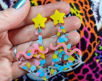 May include: A pair of clear acrylic earrings with a pink wavy line and confetti-like sprinkles. The earrings are topped with a yellow star charm.