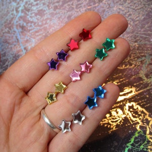 Könnte beinhalten: Eine Sammlung kleiner sternförmiger Ohrstecker in verschiedenen metallischen und reflektierenden Farben, darunter Rot, Grün, Lila, Pink, Gold, Blau und Silber. Jedes Paar Stern-Ohrstecker hat eine glatte, glänzende Oberfläche.