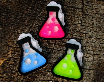 May include: Three colorful science beaker pins. Each beaker has a black outline, a black top, and white vapor. The beakers contain pink, blue, and green liquids with bubbles. The pins are set against a textured, brown wooden background.