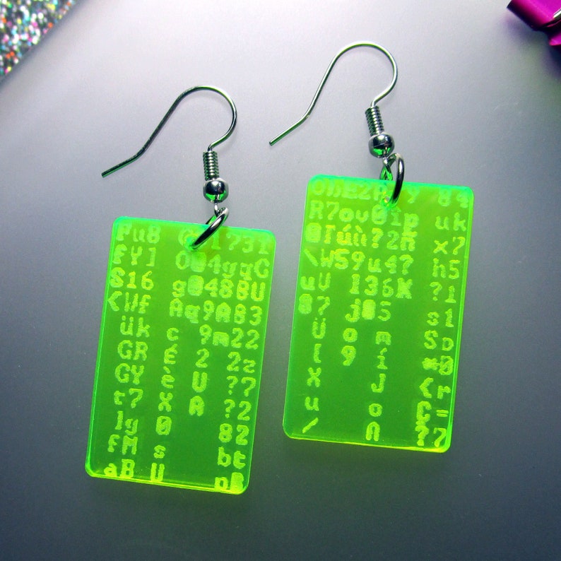 May include: A pair of green acrylic earrings with silver hooks. The earrings are rectangular and feature a pattern of letters and numbers. The text on the earrings reads: "BRZの Roubip uk T&uacute;?2r &times;? W5944? h5 87 136K ?1 u 385 31 Uom SD 1910 J <r 0 190 2 C 2 190 n 82".