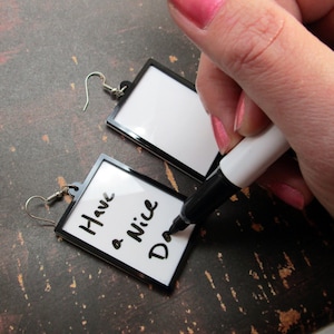 Op de afbeelding: Een paar zwarte geframed oorbellen met een wit oppervlak. Eén oorbel heeft de boodschap "Have a Nice Day" erop geschreven met een zwarte stift.