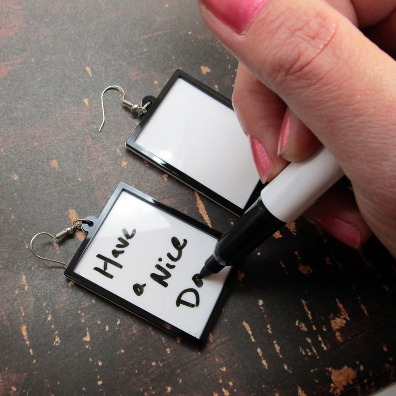 May include: A pair of black framed earrings with a white surface. One earring has the message "Have a Nice Day" written on it in black marker.