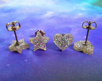 May include: Two pairs of stud earrings featuring clear resin shapes filled with iridescent glitter. One pair is shaped like five-pointed stars and the other pair is shaped like hearts. Each earring has a metallic post and a butterfly clutch backing.