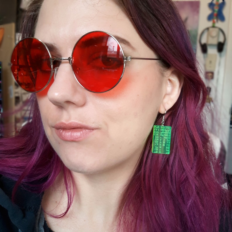 May include: A person wearing round red sunglasses and a pair of green rectangular earrings with a matrix-like code printed on them. The earrings are hanging from silver hooks.