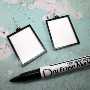 May include: Two black framed blank white rectangular earrings with silver hooks. A black dry erase marker with the text "FOR WHITEBOARD Dry Erase Mark" is in the foreground.