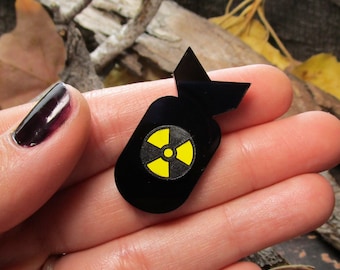 May include: A black bomb-shaped pin featuring a yellow and black radiation hazard symbol centered on the body. The pin has a smooth, glossy finish and a distinct tail fin design at the top, creating a sharp contrast between the black and yellow colors.