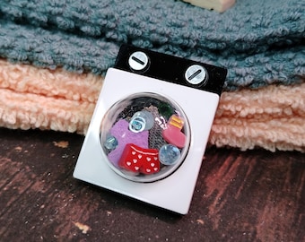 May include: A miniature white washing machine with a clear plastic window showing colorful clothes and other items inside. The washing machine is sitting on top of a stack of towels.