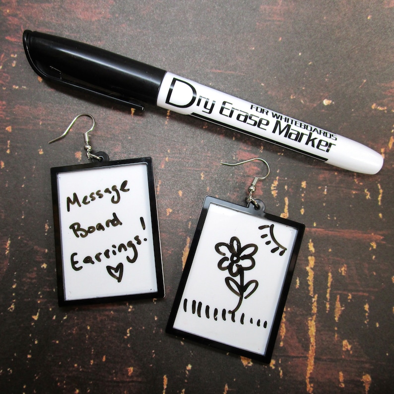 May include: Two black framed dry erase message board earrings. One earring has the text "Message Board Earrings!" written on it with a heart. The other earring has a simple drawing of a flower with a sun above it.