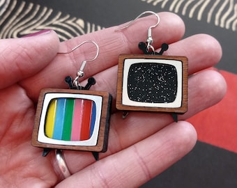 May include: A pair of wooden television-shaped earrings. One earring has a black and white screen with glitter, the other has a rainbow screen.
