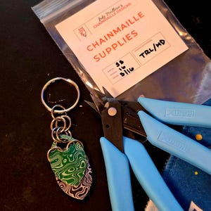 Op de afbeelding: Een sleutelhanger met een groen en zwart patroon hartvormige hanger, verbonden met een zilveren ring door kleine zilveren ringen. Een doorzichtige zak met de tekst "CHAINMAILLE SUPPLIES" en blauwe draadknippers zijn ook zichtbaar.