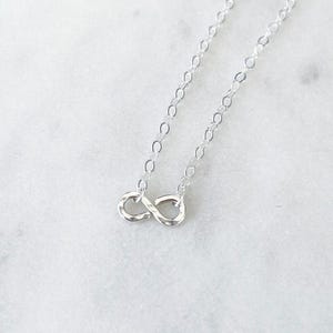 May include: A silver necklace with an infinity symbol pendant. The chain is fine, and the pendant has a polished finish. The necklace is presented on a white marble surface, with a crystal formation in the background. The necklace is a delicate piece.