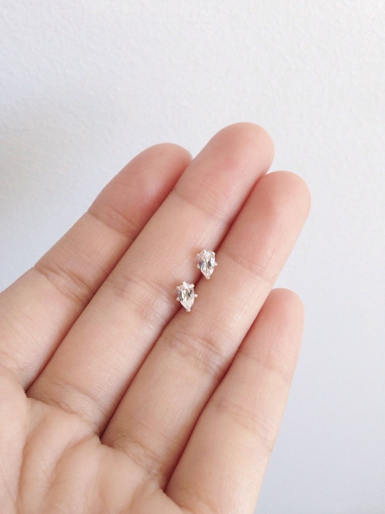 May include: A pair of small, clear, pear-shaped stud earrings. The earrings are set on a light background. The earrings are likely made of a clear gemstone or crystal, and are set in a silver or white metal.