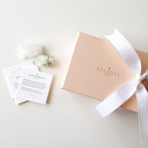 May include: A blush pink gift box tied with a white ribbon, with the brand name "SHAZOEY sydney" printed on the front. Several white cards with text and two small crystals are also present, suggesting jewelry or a similar product.