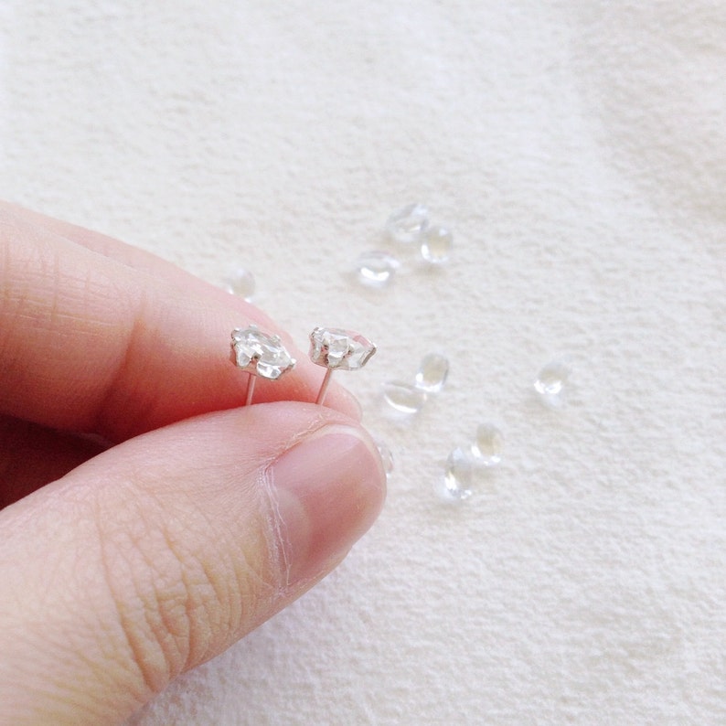 May include: A pair of silver stud earrings with clear, faceted gemstone settings. The earrings are held against a soft, white fabric background, with several loose gemstones scattered around.