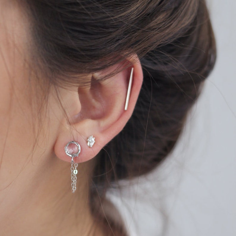 May include: A close-up of an ear adorned with multiple silver earrings. The ear features a long, straight bar earring, a small stud, and a dangling earring with a circular design and chain. The earrings are set against a blurred background.