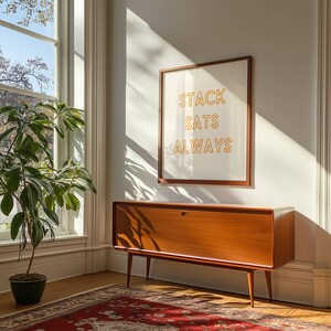 May include: A mid-century modern wooden cabinet with a single door and a light wood finish. The cabinet is sitting on a rug in front of a white wall with a framed print that reads "STACK SATS ALWAYS" in orange letters.
