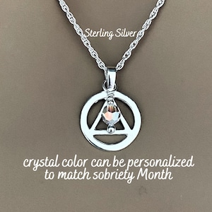 May include: Sterling silver necklace with a circle pendant featuring a triangle and a crystal. The text "Sterling Silver" is visible above the pendant. The text "crystal color can be personalized to match sobriety month" is visible below the pendant.