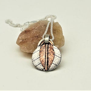 May include: A silver chain necklace with a pendant featuring a copper leaf design. The leaf is layered on top of a silver leaf with a textured surface.