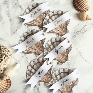 May include: Place cards shaped like seashells, each with a white banner displaying names in elegant script. The cards feature a watercolour design in brown and beige tones. Additional seashells are scattered on a marble surface around the cards.