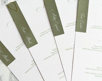 May include: A collection of white textured paper menus with olive green accents. Each menu is held together with a gold paperclip and features a guest's name in elegant script. The menus are for a wedding or special event.