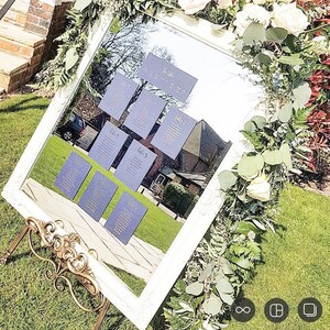 May include: A white framed mirror with a gold stand, reflecting a wedding seating chart with purple cards and gold lettering. The mirror is decorated with greenery and white flowers.