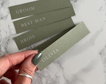 Puede incluir: Cuatro tarjetas de lugar verdes con texto blanco. Las tarjetas dicen "Groom", "Best Man", "Bride" y "Olivia".
