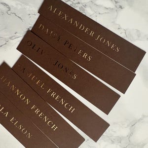 以下が含まれることがあります： 金色の文字が入った茶色の長方形の名刺。Alexander Jones、Daisy Peters、Nicola Elsonなどの名前が表示されています。カードは白とグレーの大理石の表面に配置されています。