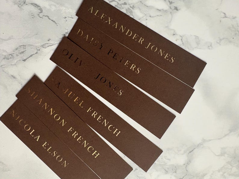 以下が含まれることがあります： 金色の文字が入った茶色の長方形の名刺。Alexander Jones、Daisy Peters、Nicola Elsonなどの名前が表示されています。カードは白とグレーの大理石の表面に配置されています。