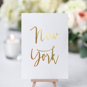 May include: A white card with gold lettering on a small wooden easel. The card says "New York" in a cursive font.