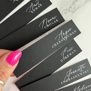 May include: Black place cards with white calligraphy names, including Darren Chambers, Amit Kumar, Neomi Cholak, Angie Chernyw Chan, Eric Dykeman, and Jennette Andrashewski. The cards are held by a hand with pink nail polish.