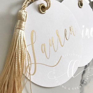 May include: White circular tags with gold foil lettering and a gold tassel. The tags feature the name "Lauren" in elegant script. The tags are for wedding and event stationery.