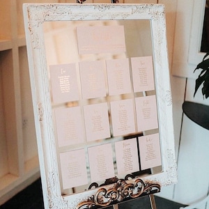 May include: A white ornate mirror frame with a gold decorative stand. The mirror is displaying a seating chart for a wedding reception. The seating chart is made up of small cards with names and table assignments written in gold lettering.