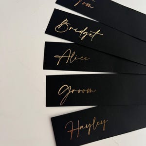 May include: Black rectangular place cards with gold foil lettering. The cards display names such as "Tom", "Bridget", "Alice", "Groom", and "Hayley". Suitable for weddings or special events.